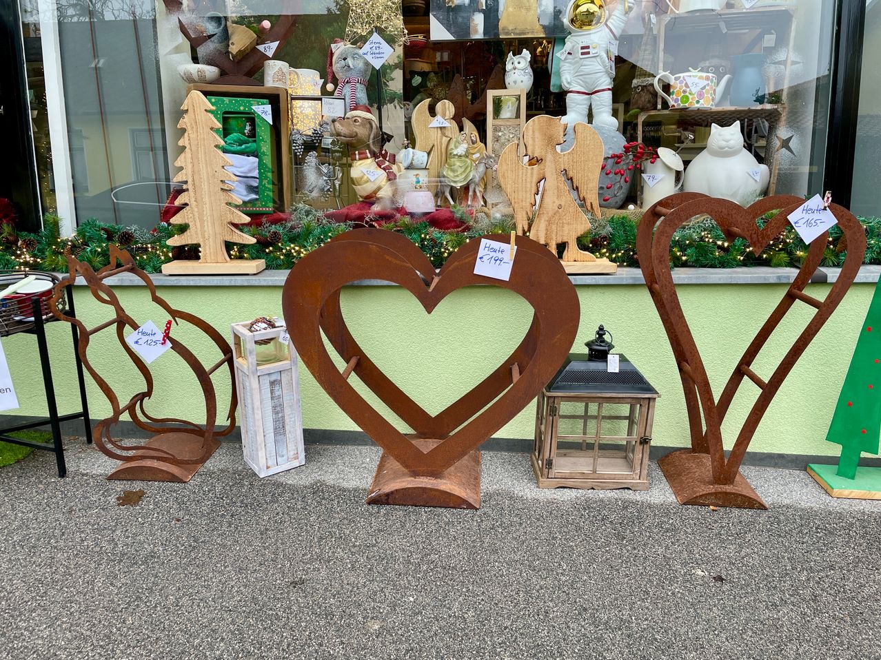 In einem Schaufenster sind mehrere dekorative herzförmige Gegenstände ausgestellt, darunter ein großes Metallherz und kleinere Holzherzen. Das Schaufenster ist mit Weihnachtsdekoration geschmückt, mit einem hölzernen Weihnachtsbaum und einem kleinen Engel auf dem Tresen.