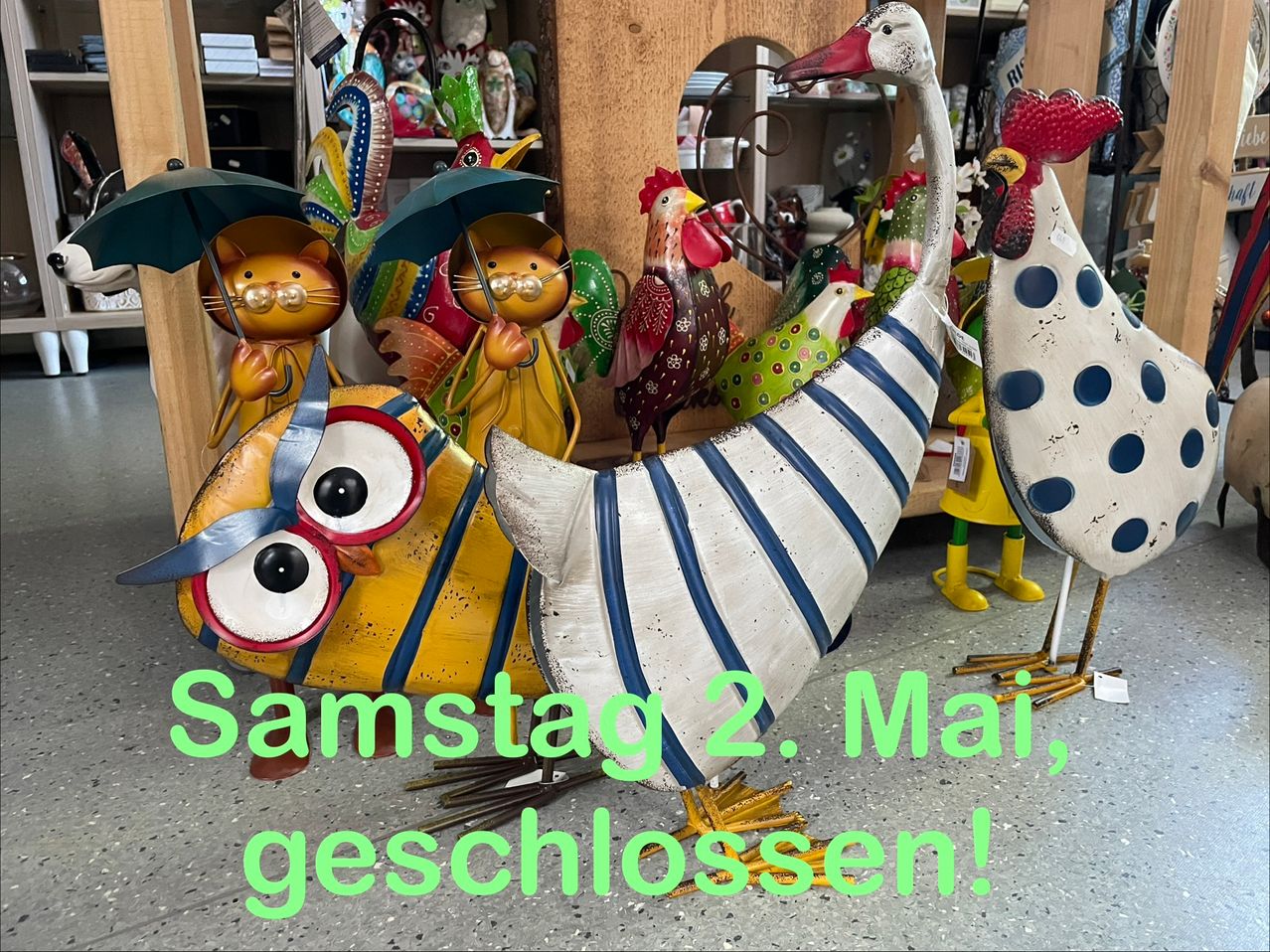 Dekorative Figuren sind in einem Geschäft ausgestellt, einschließlich Hühner, Eulen und Katzen mit Regenschirmen. Der Text-Overlay lautet 'Samstag, 2. Mai, geöffnet!' in Grün.
