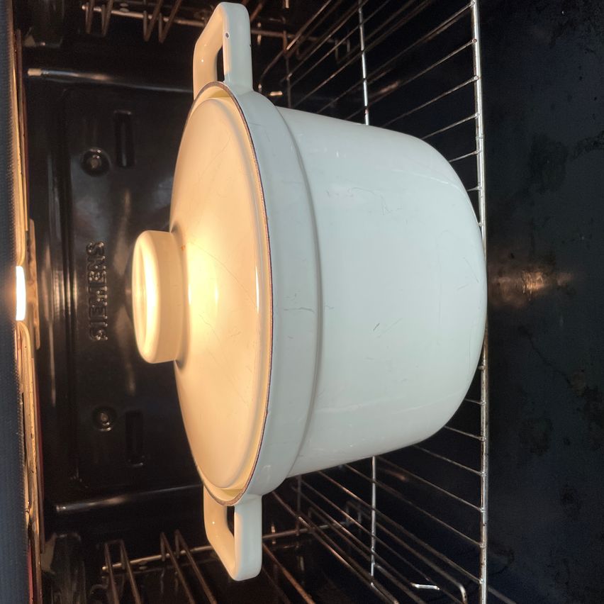 Ein weißer Keramiktopf mit Deckel befindet sich in einem Ofen. Der Ofen ist geöffnet und zeigt den Topf auf der mittleren Einschubleiste. Der Topf hat einige Abnutzungsspuren.