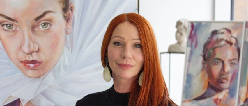 Eine Frau mit roten Haaren steht vor einem Gemälde und lächelt in die Kamera.