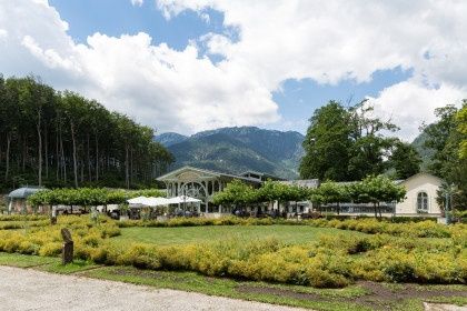 Ein ruhiger Garten mit üppigem Grün und leuchtend gelben Blumen, umgeben von Bäumen und Bergen, mit einem weißen Gebäude und Sonnenschirmen in der Ferne.