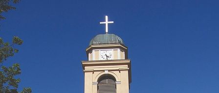 Ein hoher Turm mit einer Uhr oben und einem Symbol darunter. Er ist an ein beiges Gebäude angeschlossen.