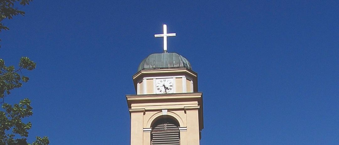 Ein hoher Turm mit einer Uhr oben und einem Symbol darunter. Er ist an ein beiges Gebäude angeschlossen.