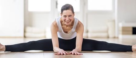 Eine Frau in einem weißen Tanktop und schwarzen Leggings macht Yoga auf dem Boden, lächelt und streckt ihre Arme.