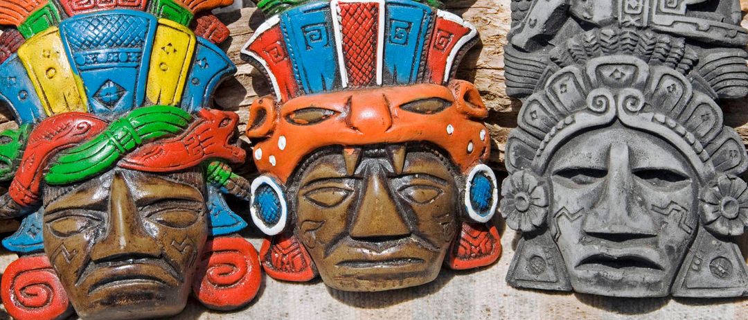 Bunte Tonmasken mit aufwendigen Mustern und Verzierungen auf einer Holzoberfläche, darunter eine mit rotem Kopftuch, blauen und weißen Ohrringen und einem blauen Auge.