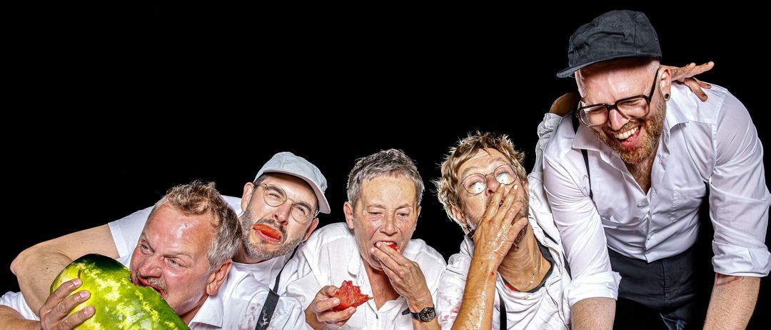 Fünf Menschen essen Wassermelonen. Einer hält eine ganze Wassermelone. Ein anderer wischt sich Wassermelonensaft vom Gesicht. Der Tisch ist mit Wassermelonensaft und Stücken bedeckt. Sie alle sehen glücklich aus.