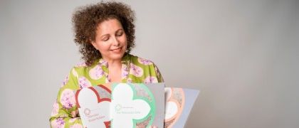 Eine Frau mit lockigem Haar und einem grünen Blumenhemd hält ein Buch mit einem grünen Cover und weißen Kleeblättern. Sie sitzt auf einem Holzschreibtisch und lächelt.