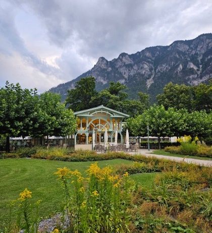 Ein Garten mit einem Pavillon und einem Berg im Hintergrund, Bäume und Gras im Vordergrund.