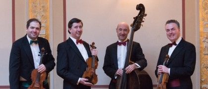 Vier Männer in schwarzen Anzügen und Fliegen stehen auf einer Bühne und halten Musikinstrumente, darunter Geigen und ein Cello. Sie posieren für ein Foto.