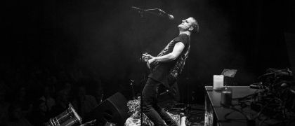 Ein Musiker spielt Gitarre auf der Bühne vor einem Publikum. Er steht neben einem Mikrofon auf einem Ständer. Die Bühne ist schwach beleuchtet.