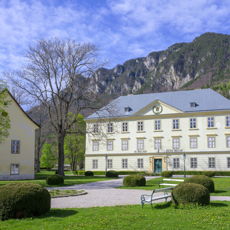 Ein großes weißes Gebäude mit grauem Dach steht vor einem Berg. Auf dem Rasen stehen zwei Bänke.
