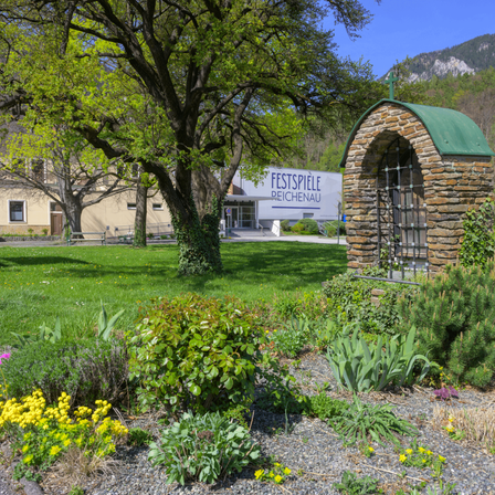 Ein Garten mit einer kleinen Kapelle auf der rechten Seite, dahinter ein Gebäude mit der Aufschrift "FESTSPIELE REICHENAU".
