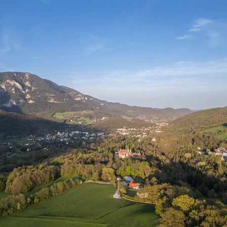 Luftaufnahme eines grünen Tals mit Bergen, Häusern und einem Schloss in der Ferne unter einem blauen Himmel.