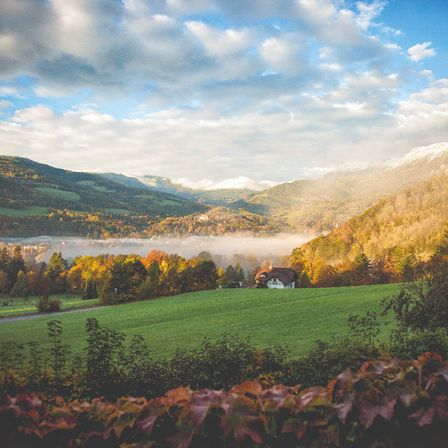 A landscape featuring a misty valley with green fields and trees in the foreground, a house with a thatched roof, and a mountain range in the background under a cloudy sky.