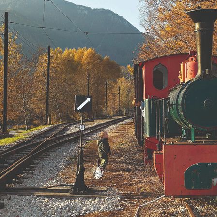 A child is running near a vintage train on the railway, with a mountain in the background.