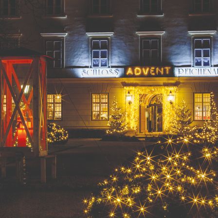 Das Schloss Advent Reichenau Hotel ist nachts wunderschön beleuchtet. Es verfügt über festliche Dekorationen, darunter eine rote Laterne und beleuchtete Weihnachtsbäume.
