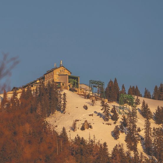 Eine Skihütte thront auf einem verschneiten Hügel, umgeben von Kiefern. Die Sonne geht im Hintergrund unter und wirft ein warmes Licht. Grüne Skilifttürme stechen gegen den verschneiten Hintergrund hervor.