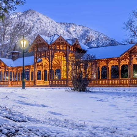 Ein Pavillon mit einem gewölbten Dach und hölzernen Geländern wird von warmen Lichtern beleuchtet, umgeben von Schnee, mit Bergen im Hintergrund.