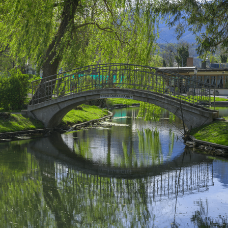 Eine Betonbrücke mit einem Metallgeländer überquert einen Fluss, umgeben von üppiger Grünfläche und einem Gebäude im Hintergrund.