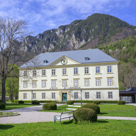 Ein großes historisches Gebäude mit grauem Dach und vielen Fenstern steht vor einem Berg. Im Vordergrund führt ein Garten mit Hecken und einem Pfad zu einer Bank.