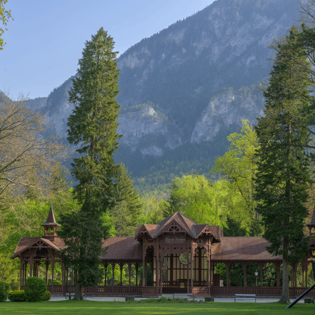 Ein Pavillon mit braunem Dach steht in einem Park, umgeben von üppigem Grün und hohen Bäumen, mit einem Berg im Hintergrund.