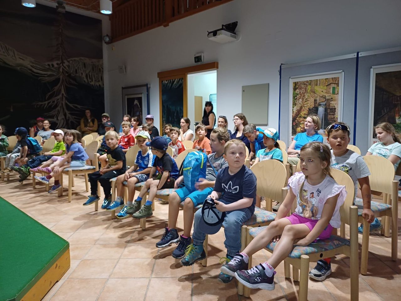 Eine Gruppe von Kindern sitzt in Stühlen in einem Raum mit gefliesten Böden und Gemälden an den Wänden. Einige tragen Mützen und Turnschuhe. Erwachsene stehen hinten.