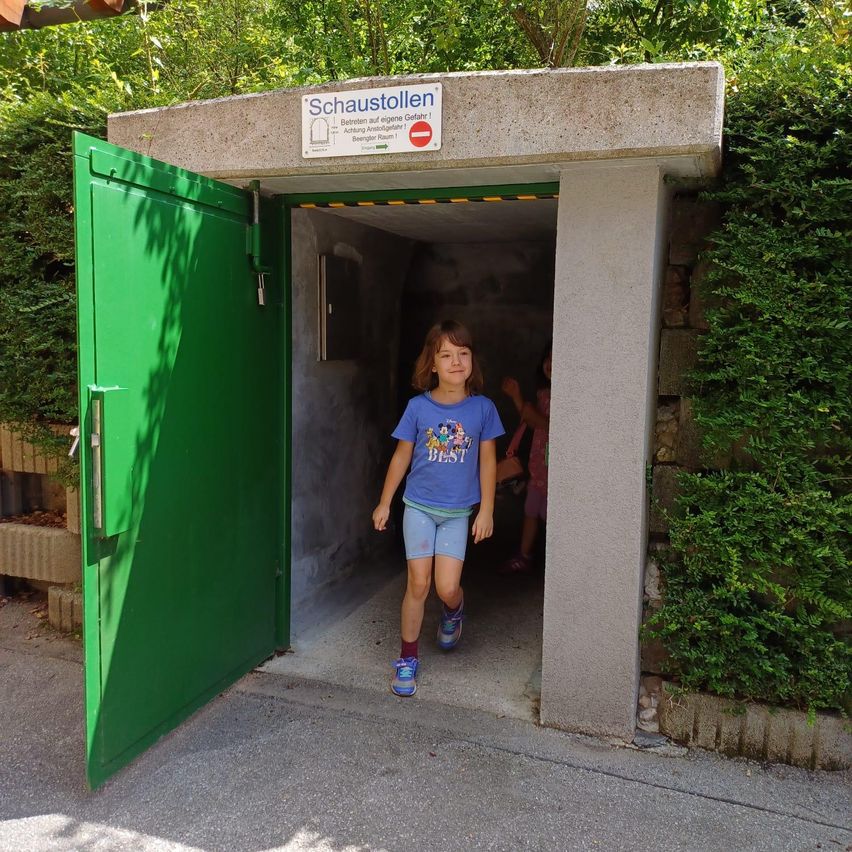 Ein kleines Mädchen verlässt einen grünen Tunnel mit einem Schild über dem Eingang, das 'Schaustellen' besagt.