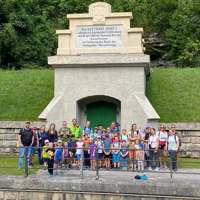 Eine Gruppe von Menschen steht vor einem Denkmal, das Kaiser Franz Josef I. gewidmet ist. Das Denkmal hat eine Plakette mit deutschem Text. Hinter dem Denkmal befindet sich eine Steinmauer und ein Grasbereich mit Bäumen.