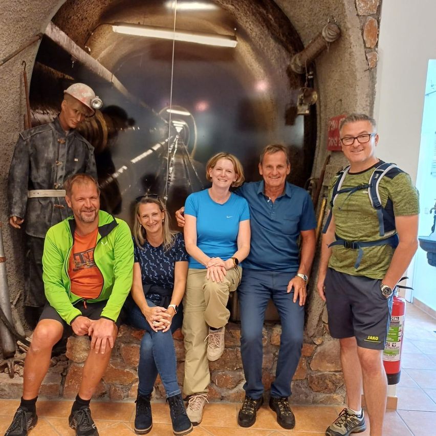 Fünf Personen posieren für ein Foto vor einer Steinmauer mit einem Tunnel-Mural. Sie tragen Freizeitkleidung, und eine Feuerwehrmann-Statue steht hinter ihnen.