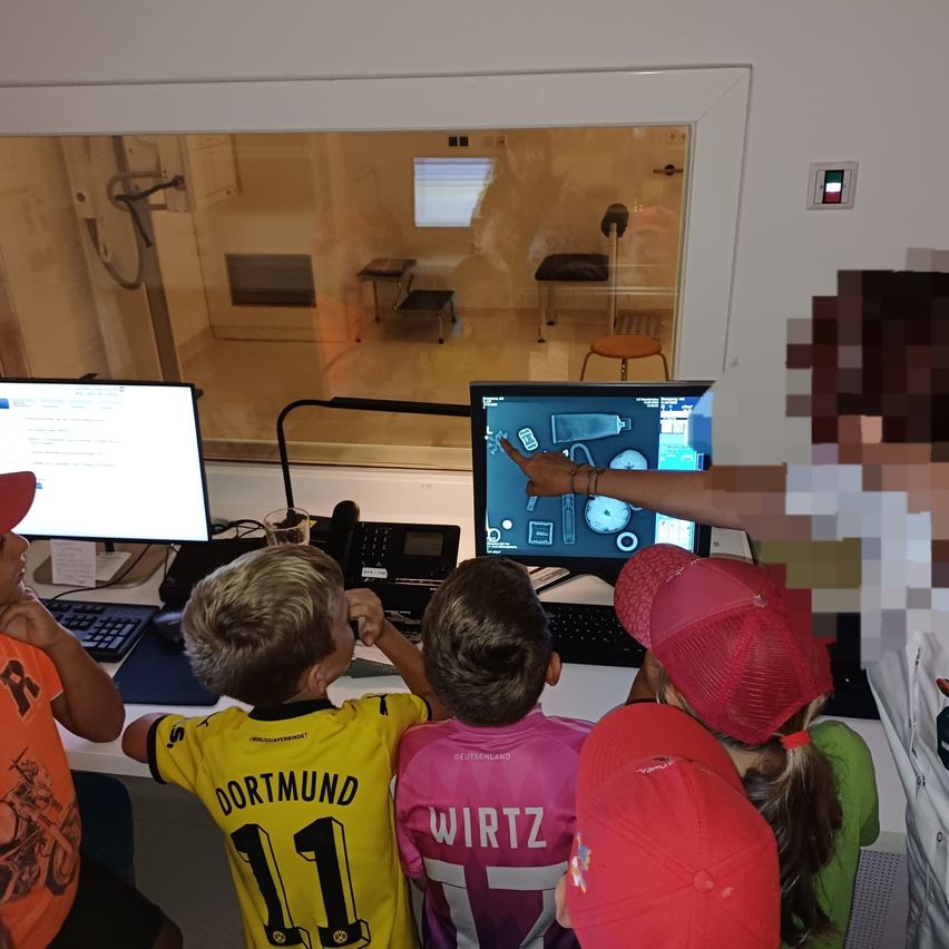 Kinder in roten Mützen beobachten einen Computerbildschirm mit einem Erwachsenen, der ein Bild zeigt. Ein Raum mit Glasfenstern, Monitoren, Tastaturen und einem Tisch.