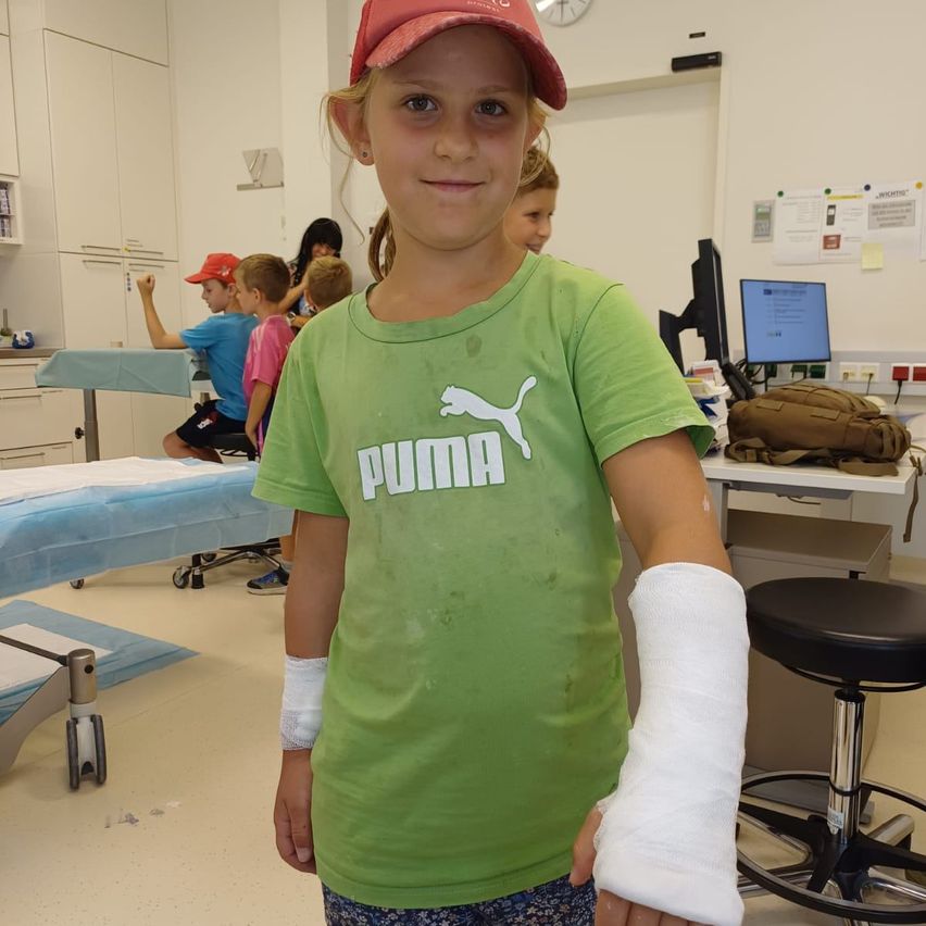 Ein junges Mädchen mit einem grünen Shirt und einer Kappe steht in einem Krankenhauszimmer mit einem gegossenen Arm. Hinter ihr sitzen andere an einem Schreibtisch mit einem Computermonitor.