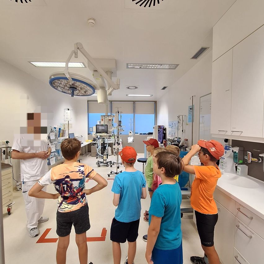 Eine Gruppe von Kindern steht in einem Krankenhauszimmer mit medizinischen Geräten und Schränken, während ein Arzt in Weiß etwas erklärt.