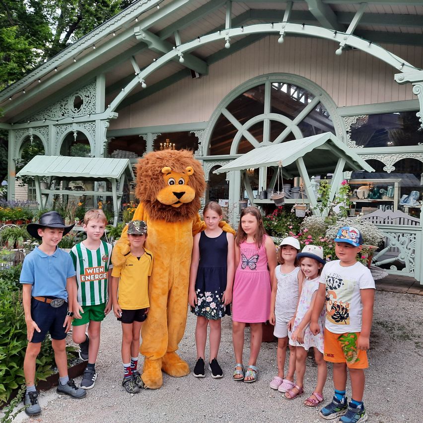 Eine Gruppe von Kindern und eine Person in einem Löwenkostüm stehen vor einem Garten. Der Garten hat eine Holzstruktur mit Markisen.