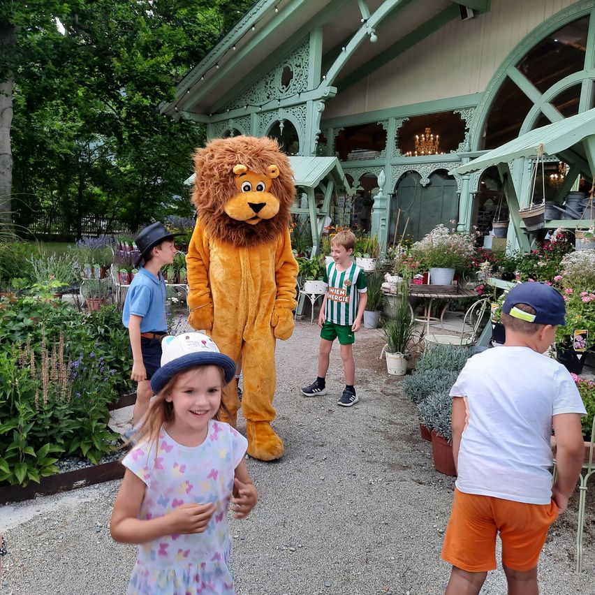 Ein Löwenmaskottchen steht in einem Garten mit Kindern, in der Nähe eines Gebäudes mit einem Pavillon und einer Kristallleuchte. Die Kinder tragen Hüte und Schuhe, und der Garten hat verschiedene Pflanzen.