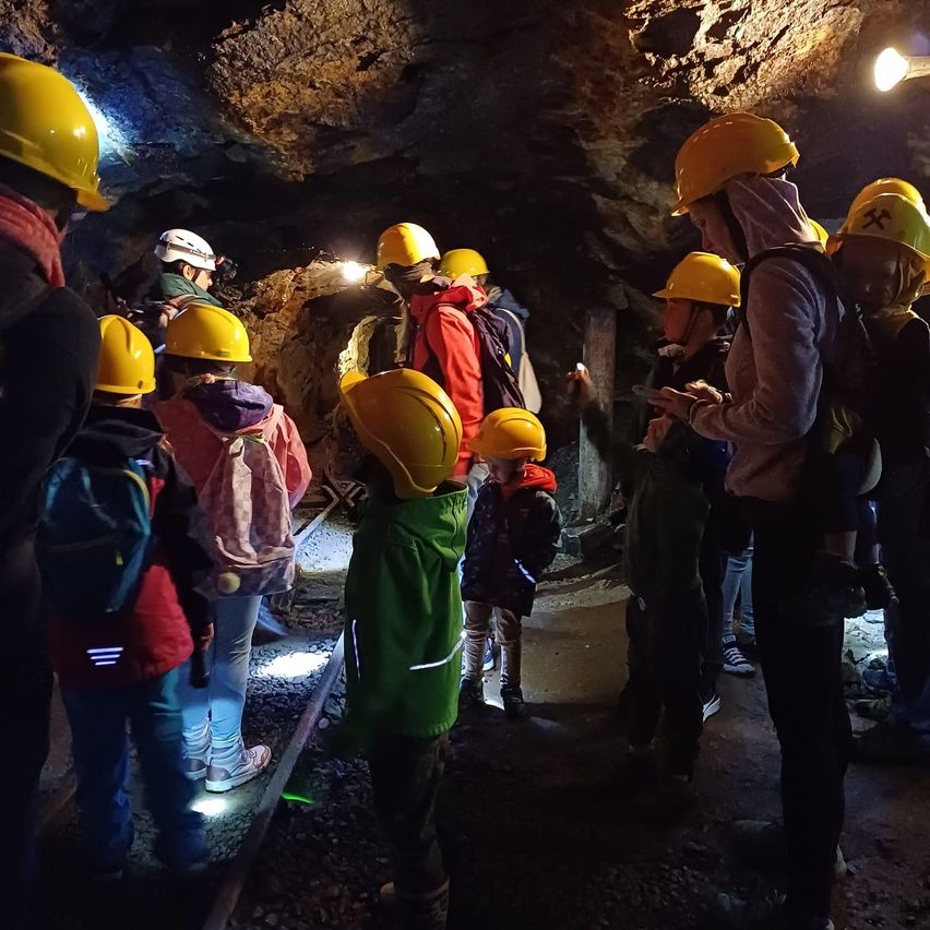 Eine Gruppe von Kindern und Erwachsenen erkundet eine Mine und trägt gelbe Helme und Rucksäcke. Sie stehen auf einem Gleis mit Steinen und Felsen.