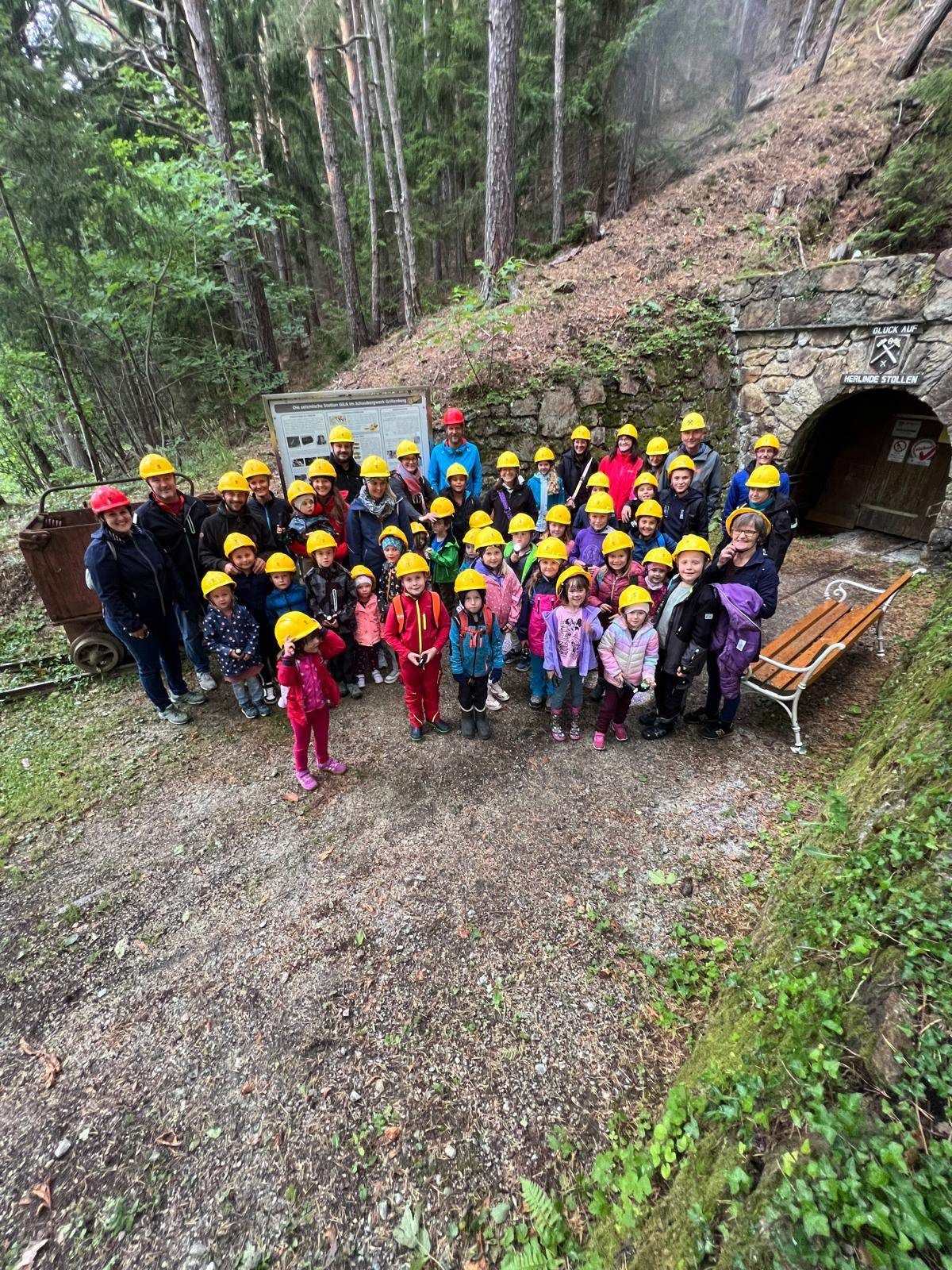 Eine Gruppe von Kindern und Erwachsenen in Helmen steht vor einem steinernen Tunneleingang mit einem Schild in einem bewaldeten Gebiet.
