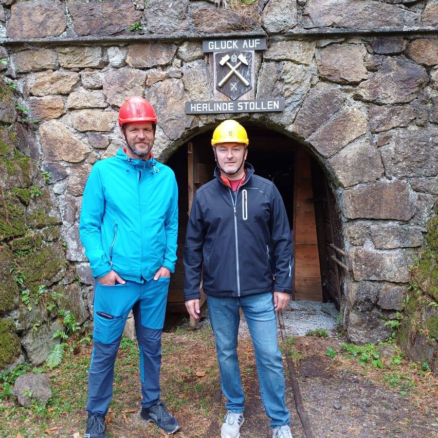 Zwei Männer in Helmen stehen vor einem steinernen Bogengang. Der Bogengang hat ein Schild, das Gluck Auf und Herlinde Stollen steht. Sie stehen auf Gras neben moosbewachsenen Felsen.