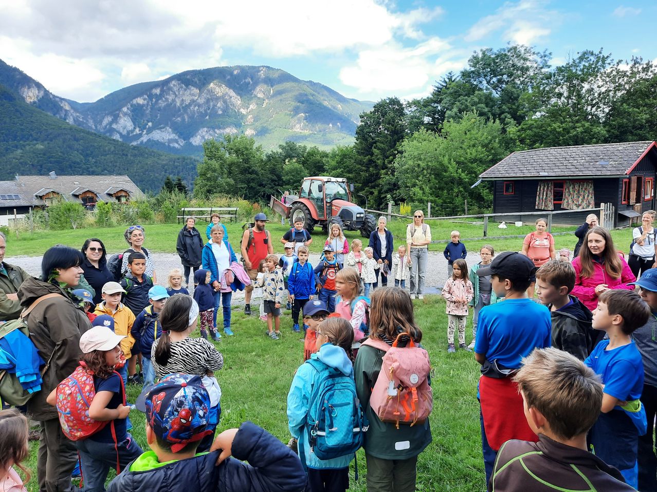 Eine Gruppe von Kindern, Erwachsenen und Lehrern versammelt sich auf einer Rasenfläche, einige mit Rucksäcken, in der Nähe eines Traktors und Häusern unter einem teilweise bewölkten Himmel.