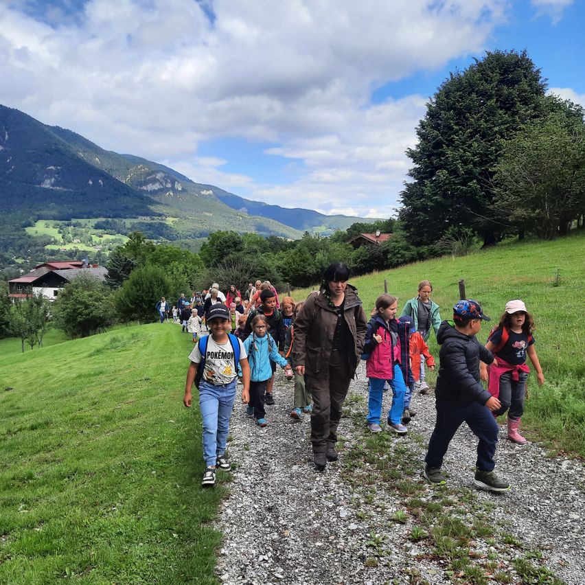 Eine Gruppe von Kindern geht einen Schotterweg in einem Gebirgsgebiet hinunter. Dahinter führt eine Frau die Gruppe an. Im Hintergrund sind Häuser und Berge zu sehen.