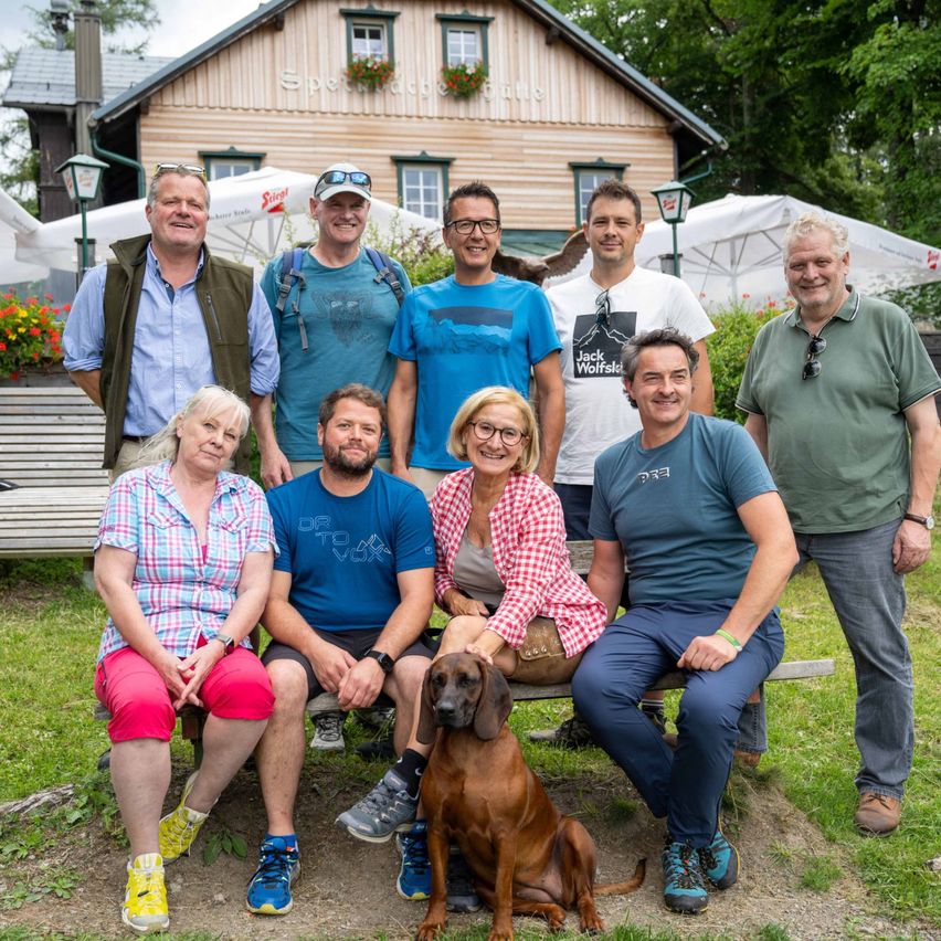Eine Gruppe von Menschen posiert für ein Foto vor einem Chalet mit Blumen, in Freizeitkleidung, und einer hält einen Hund.