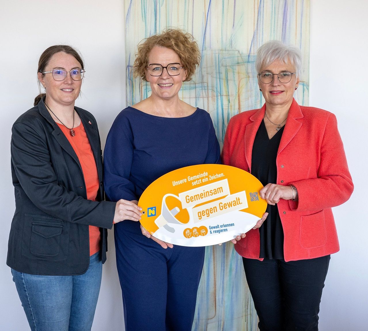 Drei Frauen stehen eng beieinander und halten ein großes gelbes Schild. Auf dem Schild steht in fettgrünen Buchstaben 'Gemeinsam gegen Gewalt'. Die Frauen tragen eine Brille und formale Kleidung. Hinter ihnen befindet sich ein abstraktes Bild an der Wand.