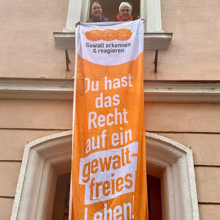 Zwei Frauen halten ein orangefarbenes Banner mit der Aufschrift 'Du hast das Recht auf ein gewaltfreies Leben' und stehen in einem Fenster.