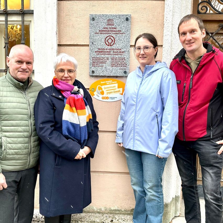 Vier Menschen stehen vor einer Wand mit einer Plakette. Sie lächeln und scheinen für ein Foto zu posieren.
