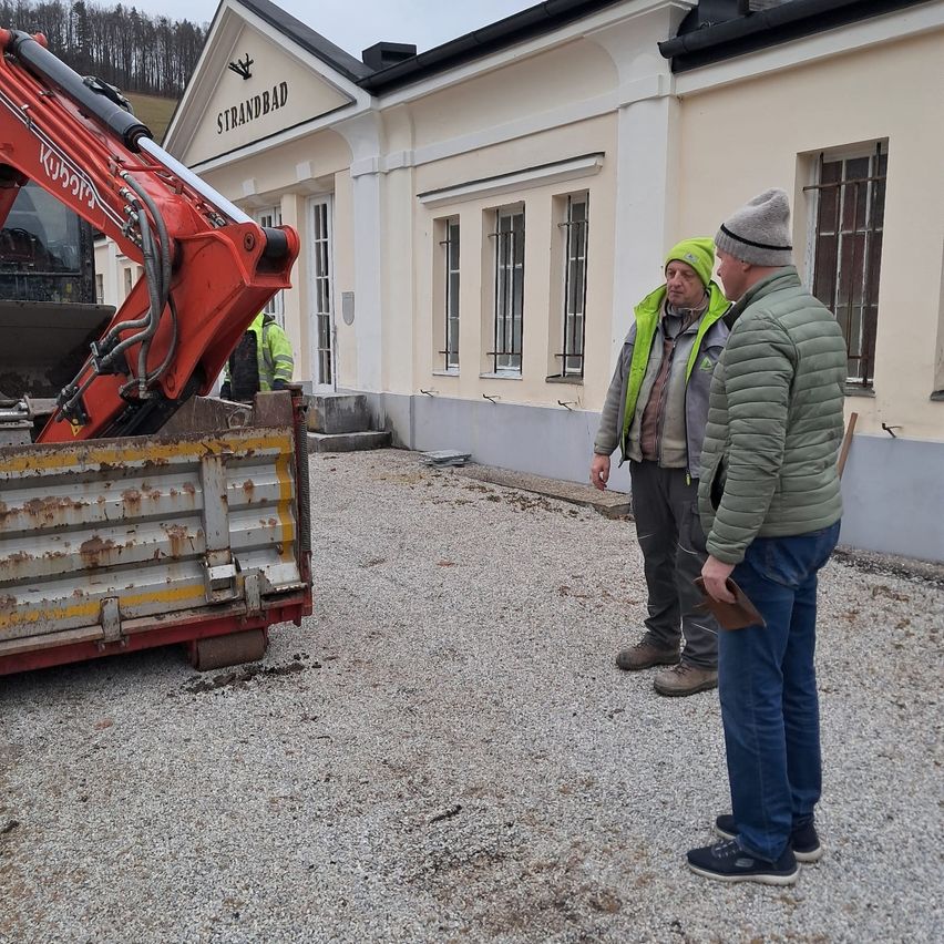 Zwei Männer stehen vor einem Gebäude mit dem Namen 'Strandbad'. Ein Mann trägt eine Warnweste und spricht. Ein Bagger steht in der Nähe.