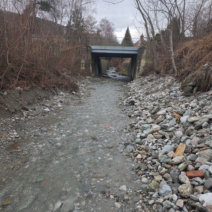 Ein kleiner Fluss mit einer Brücke darüber, umgeben von Felsen und kahlen Bäumen. Der Fluss fließt unter der Brücke hindurch.