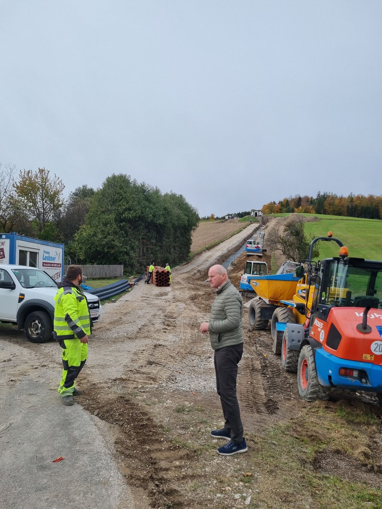 Zwei Männer stehen auf einer unbefestigten Straße, in der Nähe von Baumaschinen. Ein Mann in einer reflektierenden Jacke schaut den anderen Mann in einer grünen Jacke an. Bäume und ein Gebäude sind im Hintergrund.