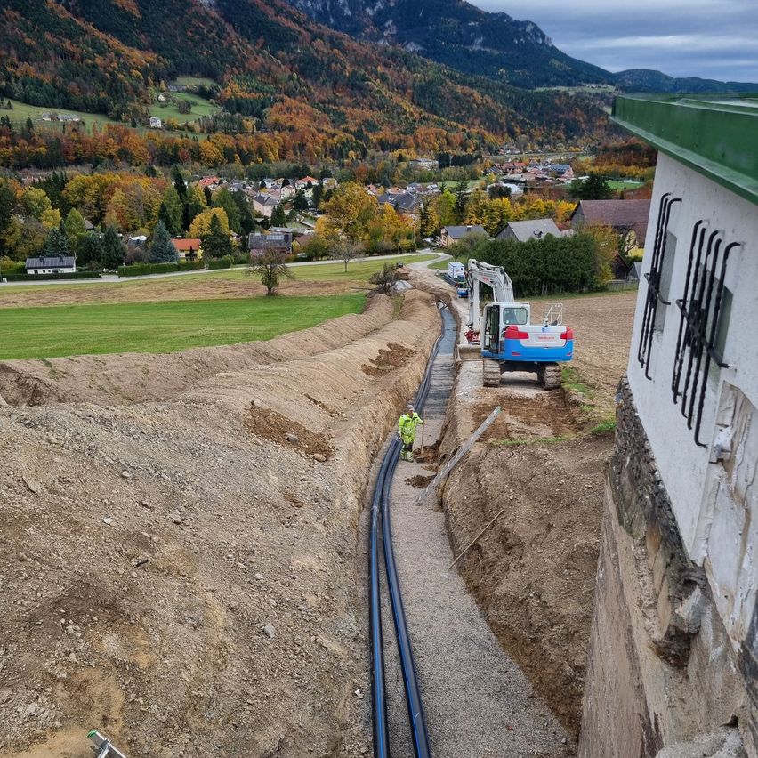 Baustelle mit einer Grube und einem Baufahrzeug. Arbeiter sind anwesend, und im Hintergrund ist eine Stadt mit Bergen zu sehen.