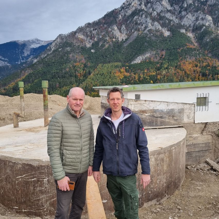 Zwei Männer stehen vor einer großen Betonstruktur, möglicherweise einem Bunker, mit einem bergigen Hintergrund.