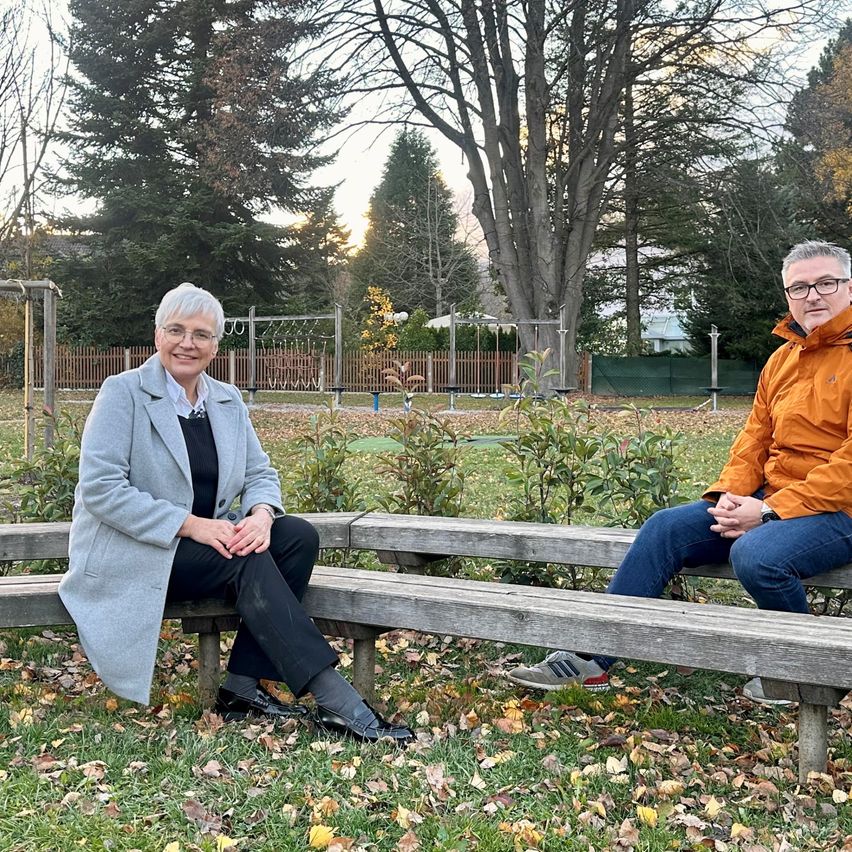 Eine Frau und ein Mann sitzen auf einer Bank in einem Park und lächeln für ein Foto. Der Park hat grünen Rasen, gefallene Blätter und Spielgeräte. Bäume umgeben das Gebiet.