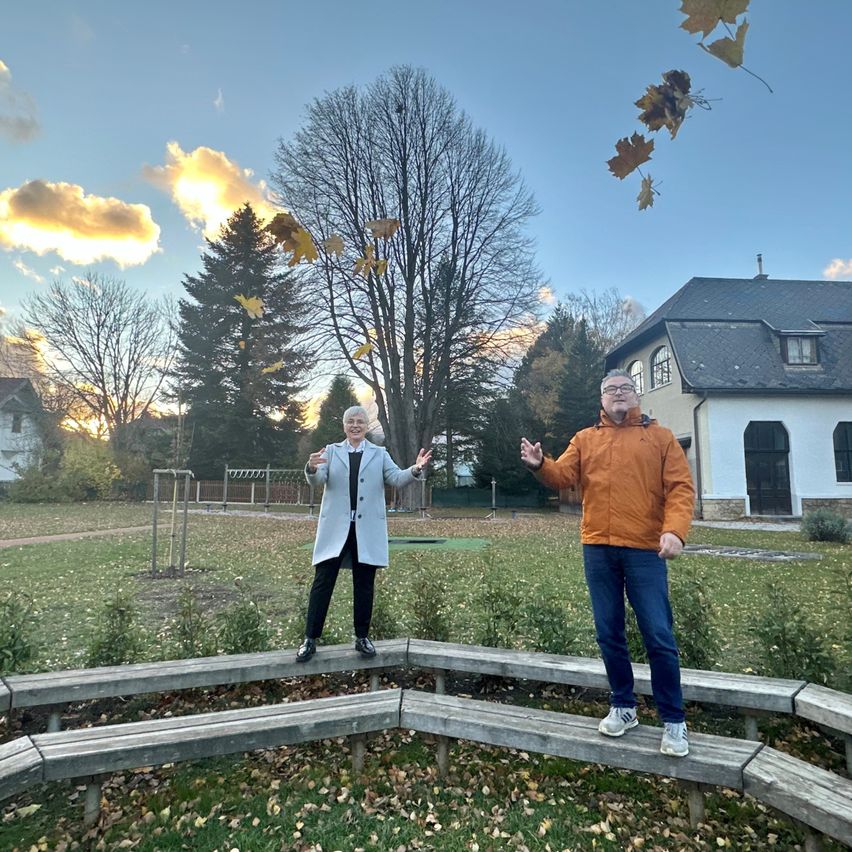 Zwei Personen stehen auf einer Holzbank in einem Park mit einem Sonnenuntergang im Hintergrund. Einer von ihnen wirft Blätter. In der Ferne befindet sich ein Haus.